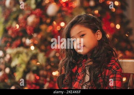 Felice giappone piccola ragazza sognatore guardando via miracolo di attesa da Santa posa vicino a noel albero. Capelli ricci neri asiatici capretto in abito rosso celebrato a plaid Foto Stock