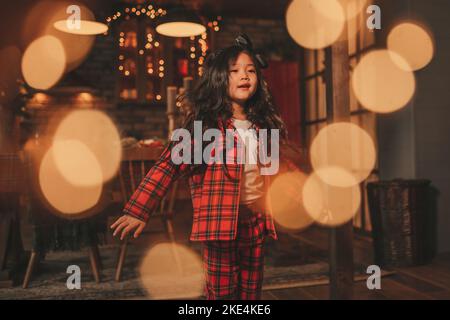 Attiva ragazza bambino giappone con elegante modello di capelli brunette lunga posa a casa in bokeh ghirlands noel. Asian piccolo fashionista in rosso plaid pajama danza Foto Stock