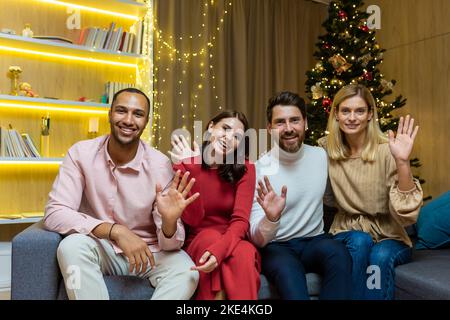Riunione online festiva. Un gruppo di giovani interracial sono seduti su un divano in un appartamento vicino ad un albero di Natale, guardando e ondeggiando la macchina fotografica, sorridendo, augurando buone vacanze di Capodanno. Foto Stock