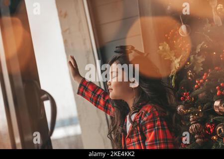 Felice giappone piccola ragazza sognatore guardando attraverso la finestra e miracolo in attesa da Santa posa vicino a noel albero. Bambino asiatico in abito rosso plaid festeggiamento Foto Stock