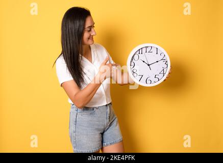 Giovane bella ragazza che tiene un grande orologio su sfondo giallo isolato molto felice ed eccitato, espressione del vincitore che celebra la vittoria urlando w Foto Stock