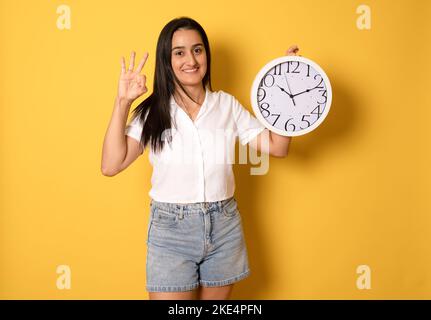 Giovane bella ragazza che tiene un grande orologio su sfondo giallo isolato molto felice ed eccitato, espressione del vincitore che celebra la vittoria urlando w Foto Stock