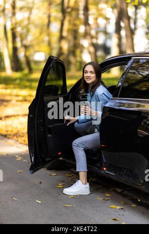 Donna attraente felice o donna di affari che indossa occhiali tenendo la tazza di caffè e uscire dalla sua auto moderna Foto Stock