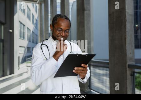 Felice e sorridente medico maturo leggere i documenti fuori clinica, l'uomo afroamericano in cappotto medico e stetoscopio e occhiali, soddisfatto del risultato del trattamento del paziente. Foto Stock