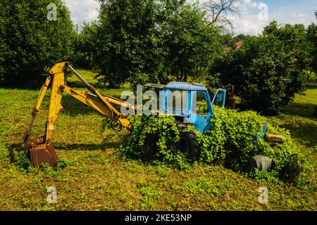 Un vecchio trattore con una benna, abbandonato da un giardino verde Foto Stock