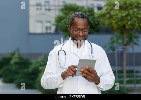 Allegro e sorridente medico senior fuori clinica utilizzando tablet computer, afro-americano indossare gli occhiali di navigazione online pagine di informazioni mediche. Foto Stock