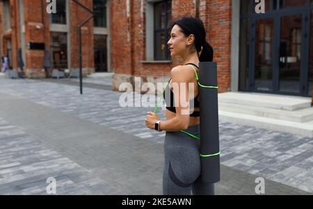Donna brunetta che porta il tappetino yoga all'aperto Foto Stock