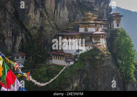 Vista panoramica della scogliera che appende il monastero buddista di Taktsang, noto anche come il Nido della Tigre, con le tradizionali bandiere di preghiera in primo piano, Paro, Bhutan Foto Stock