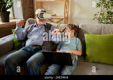 Ritratto di un uomo anziano e un ragazzino felici, nonno e nipote utilizzando gli occhiali VR seduti sul divano a casa e ridendo. Hobby, tempo libero Foto Stock