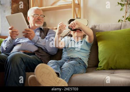 Ritratto di un uomo anziano e un ragazzino felici, nonno e nipote utilizzando gli occhiali VR seduti sul divano a casa e ridendo. Hobby, tempo libero Foto Stock