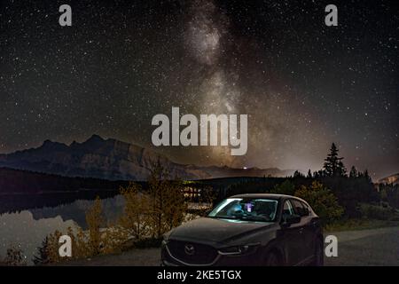 Milky Way Galaxy ambientazione dietro Mazda SUV con riflessi stellari sul lago e il SUV Hood, montagne e alberi sullo sfondo, Banff Canada Foto Stock