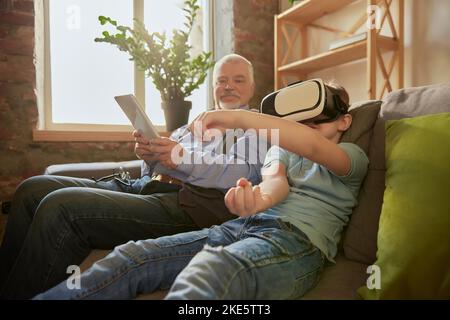 Ritratto di un uomo anziano e un ragazzino felici, nonno e nipote utilizzando gli occhiali VR seduti sul divano a casa e ridendo. Hobby, tempo libero Foto Stock