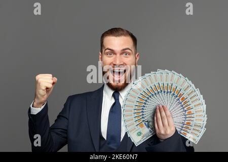 Bell'uomo d'affari felice in tuta con il ventilatore di noi dollari soldi urlando su sfondo grigio studio. Business win, divertimento e concetto di successo Foto Stock
