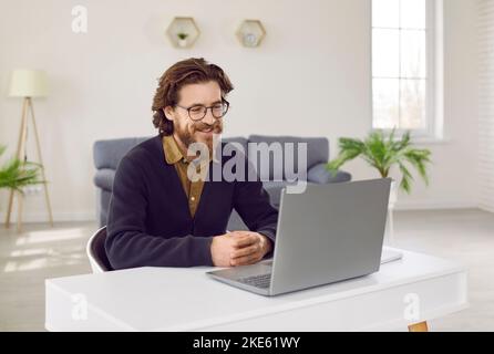 Un uomo d'affari con un sorriso guarda il notebook seduto alla scrivania in un ufficio domestico Foto Stock