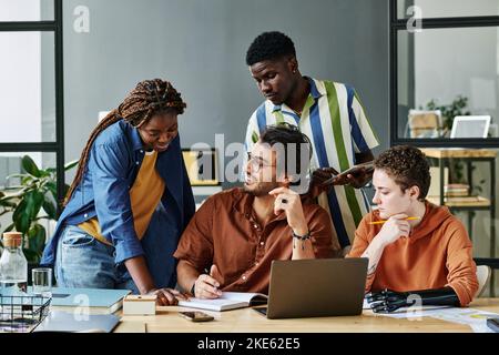 Felice giovane uomo d'affari guardando collega che indica le sue note nel copybook durante la spiegazione dei punti di lavoro tra i colleghi Foto Stock