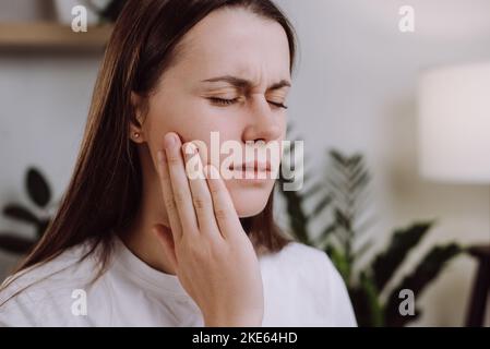 Ritratto di malsana triste giovane caucasica toccare la guancia avendo grave mal di denti a casa, bruna donna sconvolta soffre di sensazioni dolorose Foto Stock