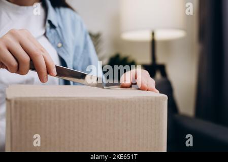 Chiudere le mani femmina utilizzando il coltello taglierina per aprire il pacco di consegna. Una giovane donna irriconoscibile seduta sul divano ha ricevuto una scatola attesa da lungo tempo con un regalo. Foto Stock