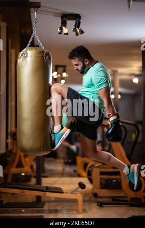 Uomo muscoloso con i guanti di boxe saltano colpendo una borsa di punzonatura con un ginocchio in una sala fitness. Foto Stock