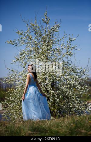 Ritratto di una ragazza tenera in un vestito lungo blu sotto un albero in fiore con un sorriso sul viso in una calda e soleggiata giornata estiva Foto Stock