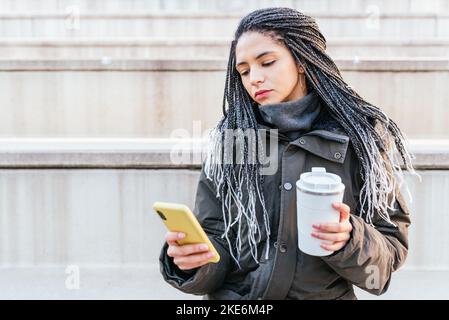 Donna ispanica focalizzata in outerwear con caffè caldo messaggi di testo sul cellulare mentre si siede sulla scala di strada della città Foto Stock