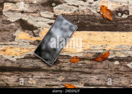 Un nuovo telefono cellulare in una custodia trasparente si trova su un vecchio tavolo con foglie autunnali cadute. Foto Stock