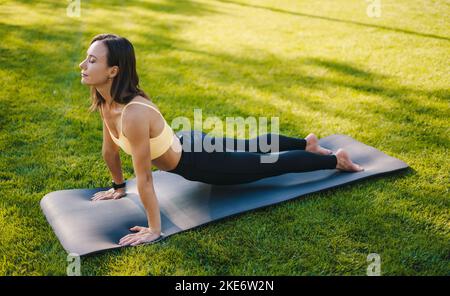 Vista laterale di una donna che pratica yoga all'aperto sdraiata su un tappetino che rende posa il cobra. Essere nel momento. Tappetino fitness, dimagrante, stretching all'aperto nel parco. Foto Stock