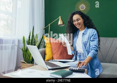 Felice donna ispanica a casa facendo lavoro d'ufficio donna calcolo del bilancio familiare e pagare bollette seduto sul divano in soggiorno utilizzando il computer portatile e calcolatrice. Foto Stock