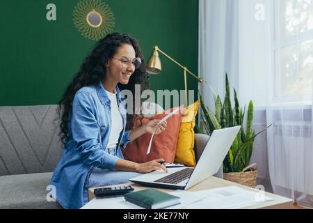 Felice donna ispanica a casa facendo lavoro d'ufficio donna calcolo del bilancio familiare e pagare bollette seduto sul divano in soggiorno utilizzando il computer portatile e calcolatrice. Foto Stock