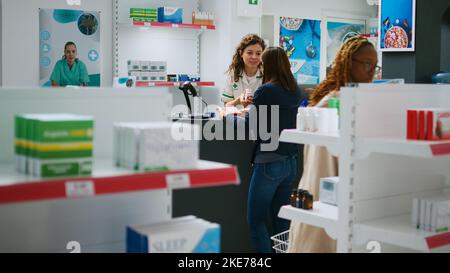 Farmacista spiegare pillole opuscolo al cliente asiatico al banco farmacia, parlando di farmaci e integratori. Lavoratore che aiuta i clienti con prodotti farmaceutici in farmacia. Foto Stock