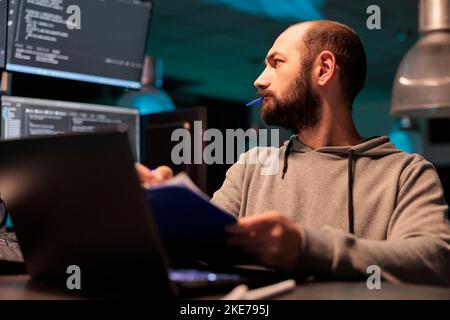 Sviluppatore web analizzando il codice sorgente con documenti e computer, prendendo appunti e lavorando allo sviluppo web. Programmatore maschio che confronta i file con i dati del server online dopo ore. Foto Stock