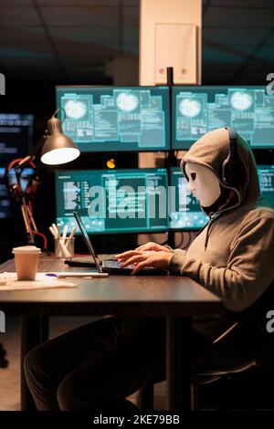 Pericoloso hacker mascherato rubare informazioni dal server di database online, hacking computer hardware sistema. Impostor con cappuccio e maschera che creano malware e violazione della sicurezza in ufficio. Foto Stock