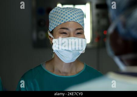 Chirurgo asiatico femminile sorridente a un collega in sala operatoria durante il funzionamento Foto Stock