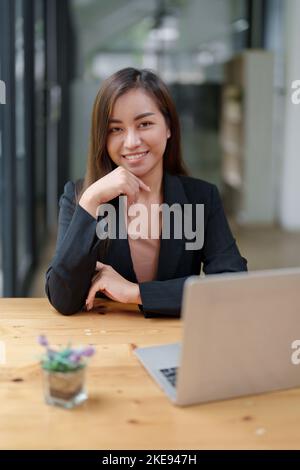 Il ritratto di una bella donna d'affari asiatica in abito formale si siede alla sua scrivania con un computer portatile in ufficio Foto Stock