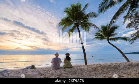 Un paio di uomini e donne seduti sulla spiaggia guardando il tramonto sulla spiaggia con sabbia bianca e palme, Bang Tao Beach Phuket Thailandia. Foto Stock