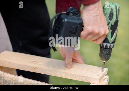 Man sta avvitando la vite nella scatola di legno con un cacciavite a batteria. Riparazione del prodotto con un cacciavite elettrico professionale. Primo piano. Foto di alta qualità Foto Stock