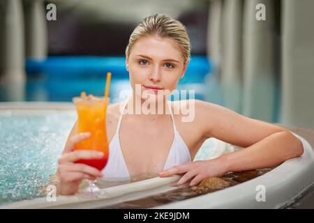 Bolle ai miei piedi e bere nella mia perfezione di mano. Ritratto di una giovane donna che beve un cocktail in piscina presso una spa. Foto Stock