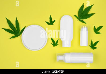 Cosmetici naturali di canapa. Tubi cosmetici in plastica bianca per crema e olio con foglie di canapa su sfondo giallo, mock up, piatto. Foto Stock