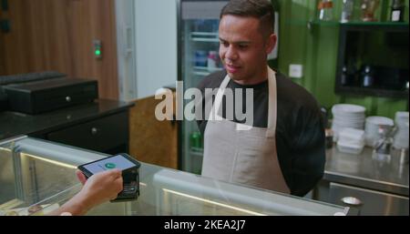 Persona che paga con il cellulare al banco del caffè. Barista che consegna la macchina ATM per il pagamento NFC senza contatto tramite smartphone. Pagamento accettato animat Foto Stock