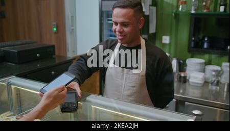 Persona che paga con il cellulare al banco del caffè. Barista che consegna la macchina ATM per il pagamento NFC senza contatto tramite smartphone. Pagamento accettato animat Foto Stock