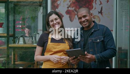 Un manager afro-americano di piccole imprese che tiene un tablet che parla con una dipendente femminile all'ingresso della caffetteria. Ritratto di due diversi staf felice Foto Stock