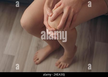 Vista dall'alto di una giovane donna irriconoscibile che tocca l'articolazione del ginocchio, avendo sensazioni dolorose seduto sul divano. Donna malsana che soffre di artrite, ossa Foto Stock