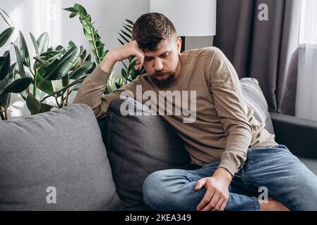 Maschio ponderato meditando sopra il problema o la difficoltà. Uomo caucasico infelice seduto sul divano pensando alla soluzione del problema, focalizzato giovane ragazzo perso su di te Foto Stock