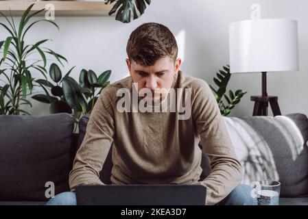 Primo piano di un giovane uomo concentrato seduto su un comodo divano a casa lavorando su un moderno gadget per laptop, focalizzato 30s maschio bearded a tavolo che naviga in wireless Foto Stock
