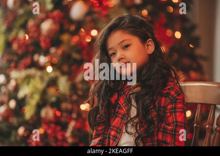 Felice giappone piccola ragazza sognatore guardando via miracolo di attesa da Santa posa vicino a noel albero. Capelli ricci neri asiatici capretto in abito rosso celebrato a plaid Foto Stock