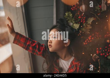 Felice giappone piccola ragazza sognatore guardando attraverso la finestra e miracolo in attesa da Santa posa vicino a noel albero. Bambino asiatico in abito rosso plaid festeggiamento Foto Stock