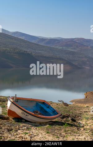 Vecchia canoa in legno, barca di colore blu, arancione e bianco arenata su terra sterrata asciutta nella diga del lago Bouzegza Keddara con le montagne nebbre riflesse. Foto Stock