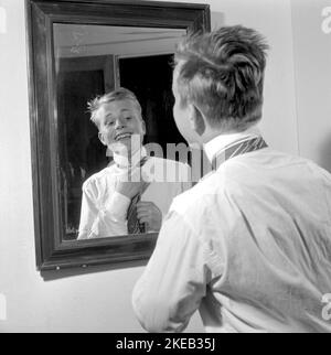 Vestirsi con la 1950s. Un giovane uomo davanti allo specchio gli node la cravatta. Ci sono varianti di legatura della cravatta tra cui Ascot, prua, bolo, cravatta con cerniera, cravat e maglia. Al momento in cui la foto è stata presa la moda e lo sguardo delle cravatte erano più sottili e sottili che i decenni precedenti. Ci sono quattro nodi principali utilizzati per il nodo cravatta: Quattro in mano nodo, Pratt nodo, mezzo Windsor nodo e Windsor nodo. Svezia 1959. Rif. Conard 4019 Foto Stock