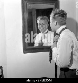 Vestirsi con la 1950s. Un giovane uomo davanti allo specchio gli node la cravatta. Ci sono varianti di legatura della cravatta tra cui Ascot, prua, bolo, cravatta con cerniera, cravat e maglia. Al momento in cui la foto è stata presa la moda e lo sguardo delle cravatte erano più sottili e sottili che i decenni precedenti. Ci sono quattro nodi principali utilizzati per il nodo cravatta: Quattro in mano nodo, Pratt nodo, mezzo Windsor nodo e Windsor nodo. Svezia 1959. Rif. Conard 4019 Foto Stock