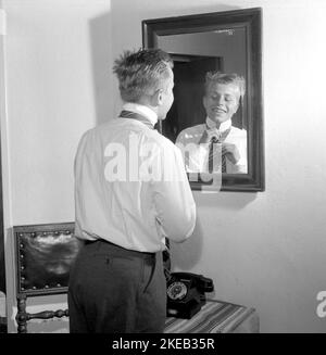 Vestirsi con la 1950s. Un giovane uomo davanti allo specchio gli node la cravatta. Ci sono varianti di legatura della cravatta tra cui Ascot, prua, bolo, cravatta con cerniera, cravat e maglia. Al momento in cui la foto è stata presa la moda e lo sguardo delle cravatte erano più sottili e sottili che i decenni precedenti. Ci sono quattro nodi principali utilizzati per il nodo cravatta: Quattro in mano nodo, Pratt nodo, mezzo Windsor nodo e Windsor nodo. Svezia 1959. Rif. Conard 4019 Foto Stock