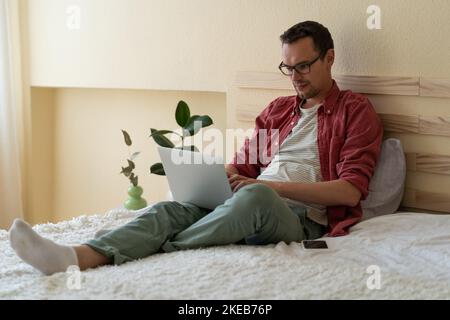 L'uomo caucasico concentrato si siede sul letto nella camera dell'hotel leggendo le notizie via internet o controllando la posta elettronica Foto Stock